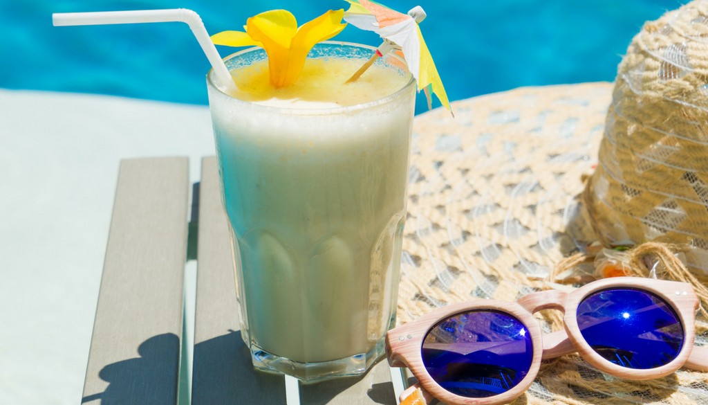 A Piña colada drink next to a straw hat and wooden glasses outside next to a pool.