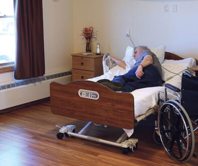 A senior sleeping in a electric bed with a wheelchair nearby.