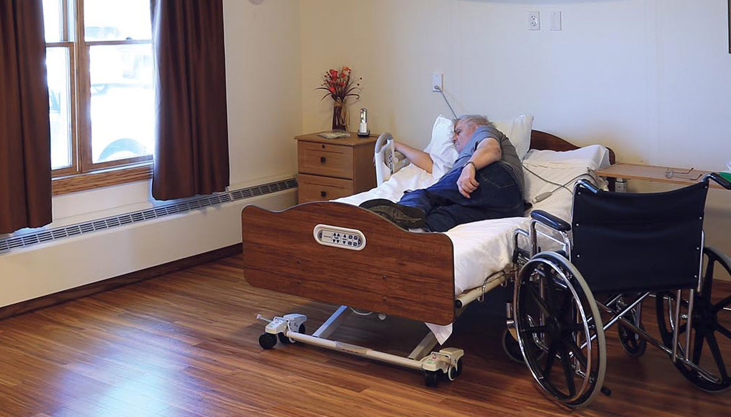 A senior sleeping in a electric bed with a wheelchair nearby.