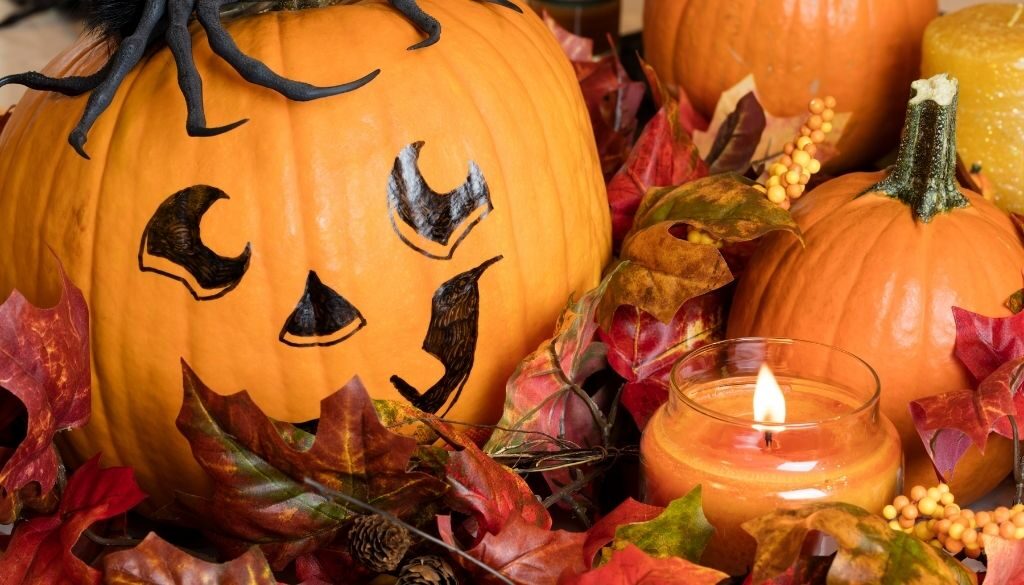 Large Halloween pumpkin with a black jack-o-lantern face painted on it surrounded by fall leaves , smaller pumpkins and a lit candle.