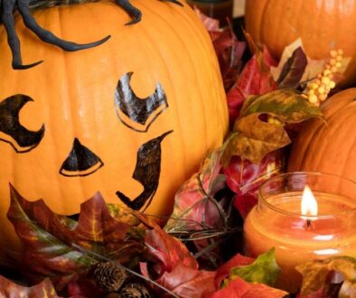 Large Halloween pumpkin with a black jack-o-lantern face painted on it surrounded by fall leaves , smaller pumpkins and a lit candle.