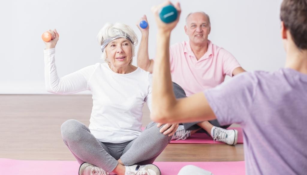 An elderly man and woman representing active seniors sitting on yoga mats exercising with weighted dumbbells.