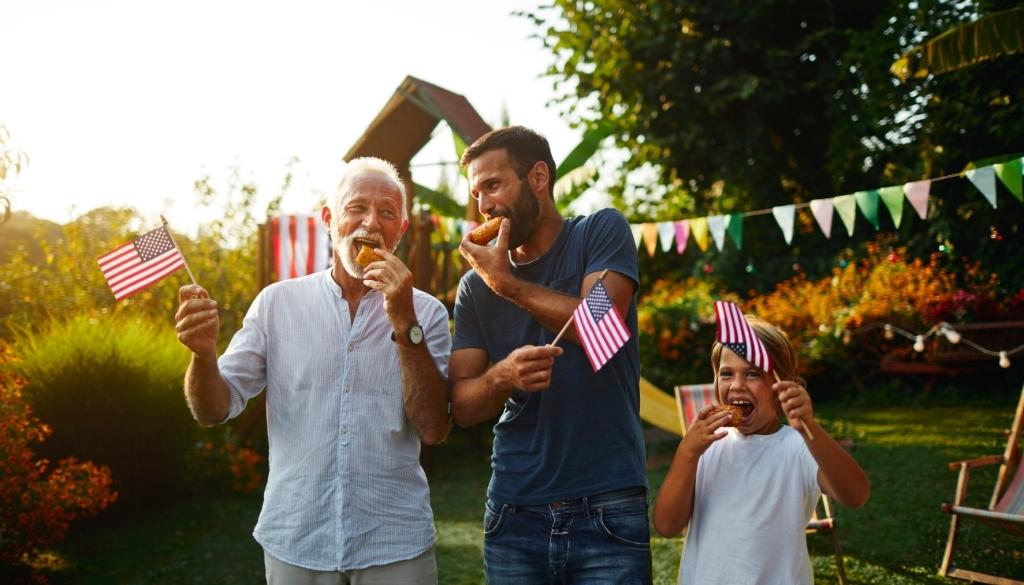 A grandpa, father, and son enjoying corn on the cob and 4th of July activities together.