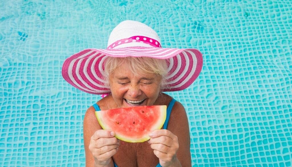 An older woman in a blue pool eating watermelon wearing a blue swimsuit and pink sun hat.
