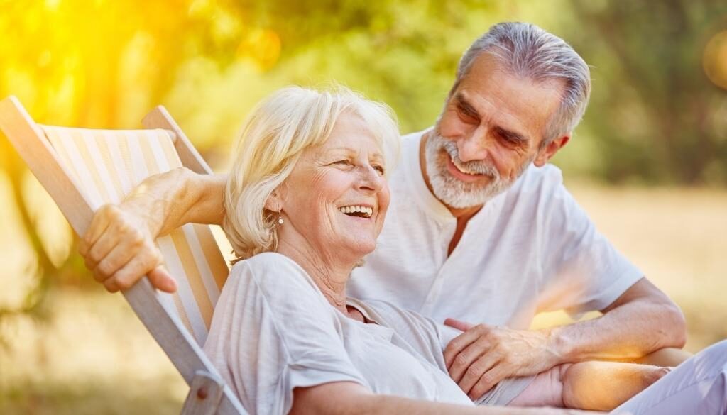 An older man and woman laughing and smiling while sitting with the sun shining on their skin.