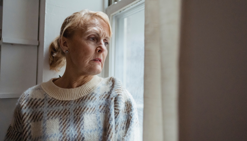 Elderly woman looking out gloomy window on a rainy day. The Neighbors of Dunn County Seasonal Affective Disorder (SAD) in elderly adults.