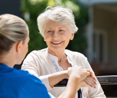Elderly woman in a wheelchair smiles while a nurse sits next to her.