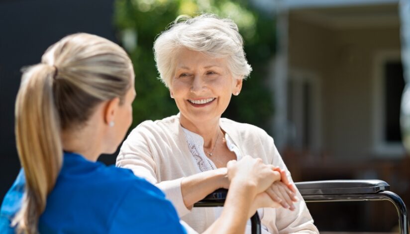 Elderly woman in a wheelchair smiles while a nurse sits next to her.