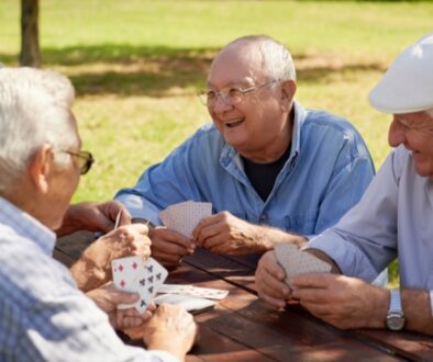 A group of elderly men laughing and smiling while playing a game of cards in an outdoor area.