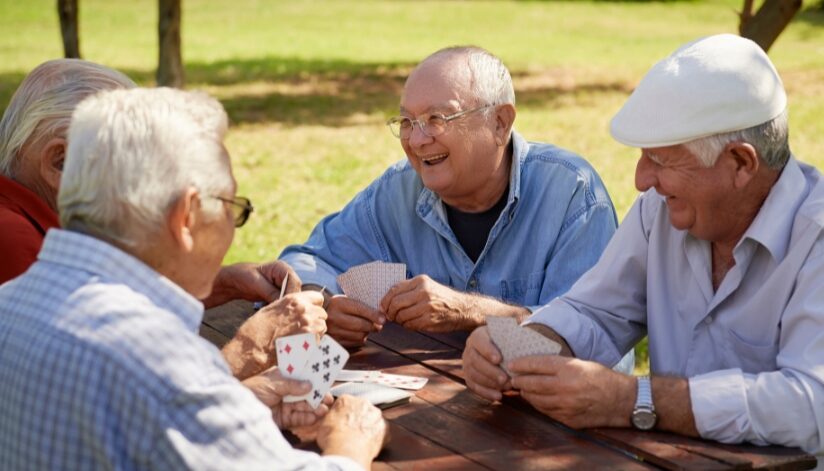 A group of elderly men laughing and smiling while playing a game of cards in an outdoor area.
