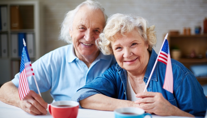 Two older adults, one male and one female, hold an American flag and look into the camera.