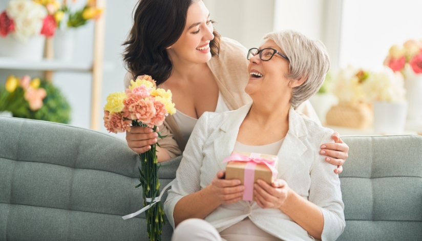 Older woman looks up from where she is sitting on a couch to her adult daughter holding flowers to give her and smiling at her.