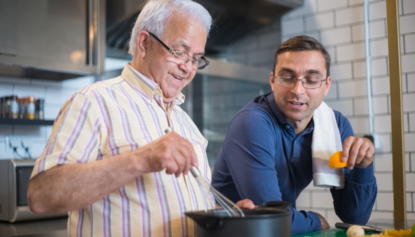 Older father and his grown son stand over a pot and cook together.