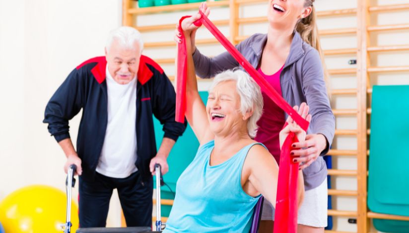 Older woman using a resistance band during physical therapy with assistance from a therapist, while an older man exercises nearby.