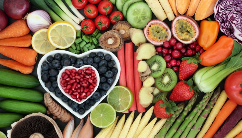 Fresh fruits and vegetables arranged around a heart-shaped bowl filled with blueberries and pomegranate seeds.